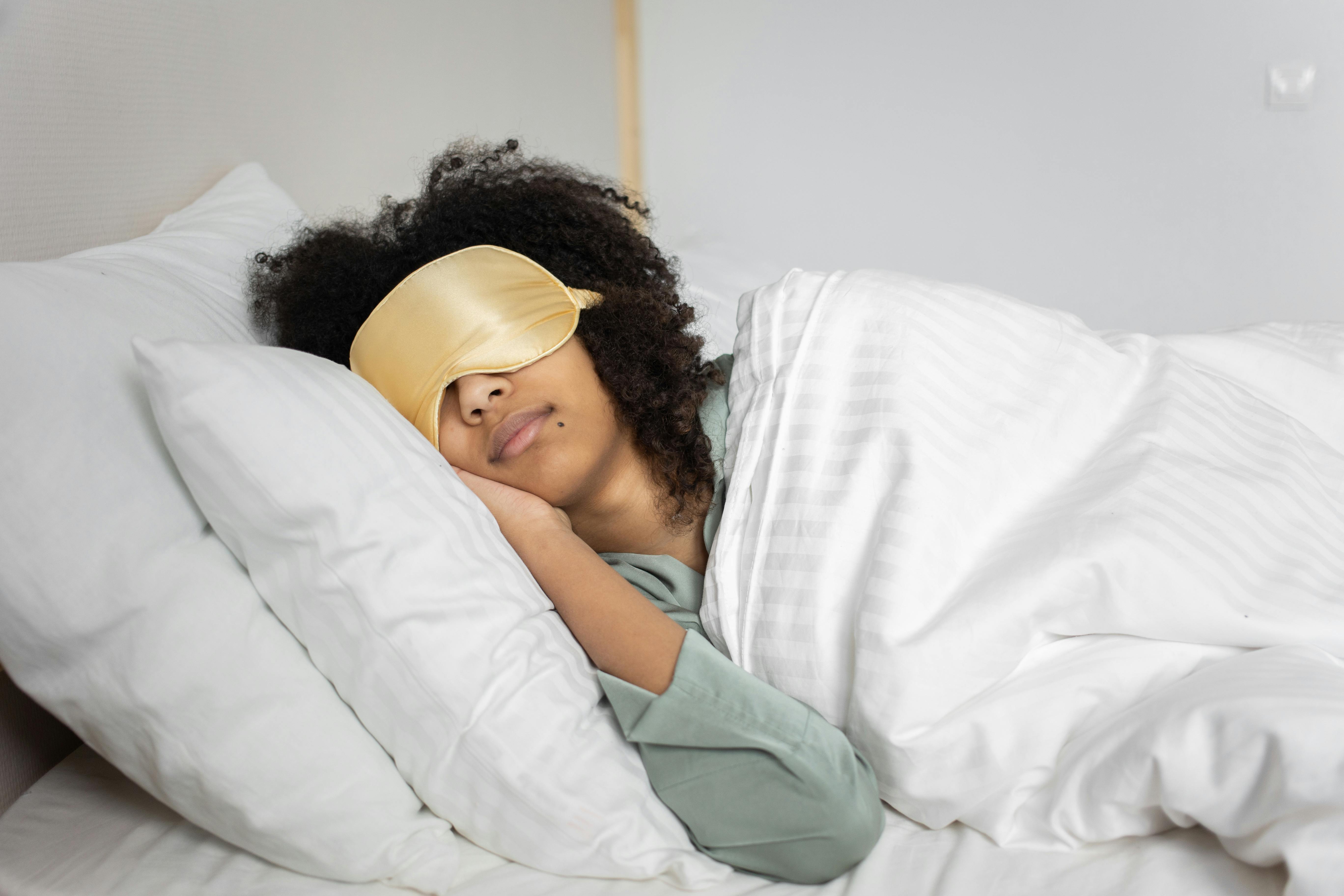 Woman with curly hair and light gold sleeping mask laying on fluffy white pillows with fluffy white comforter.